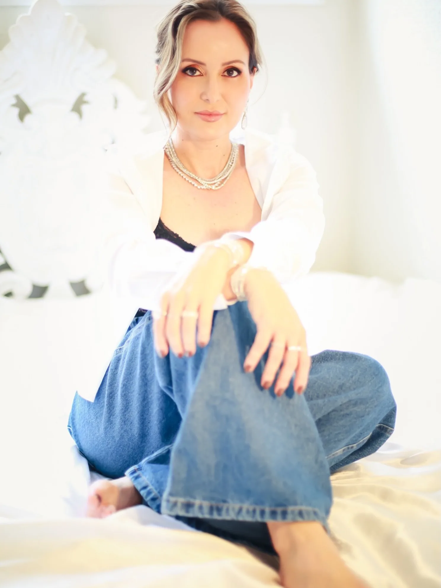 Mulher sentada na cama, com camisa branca e calça jeans