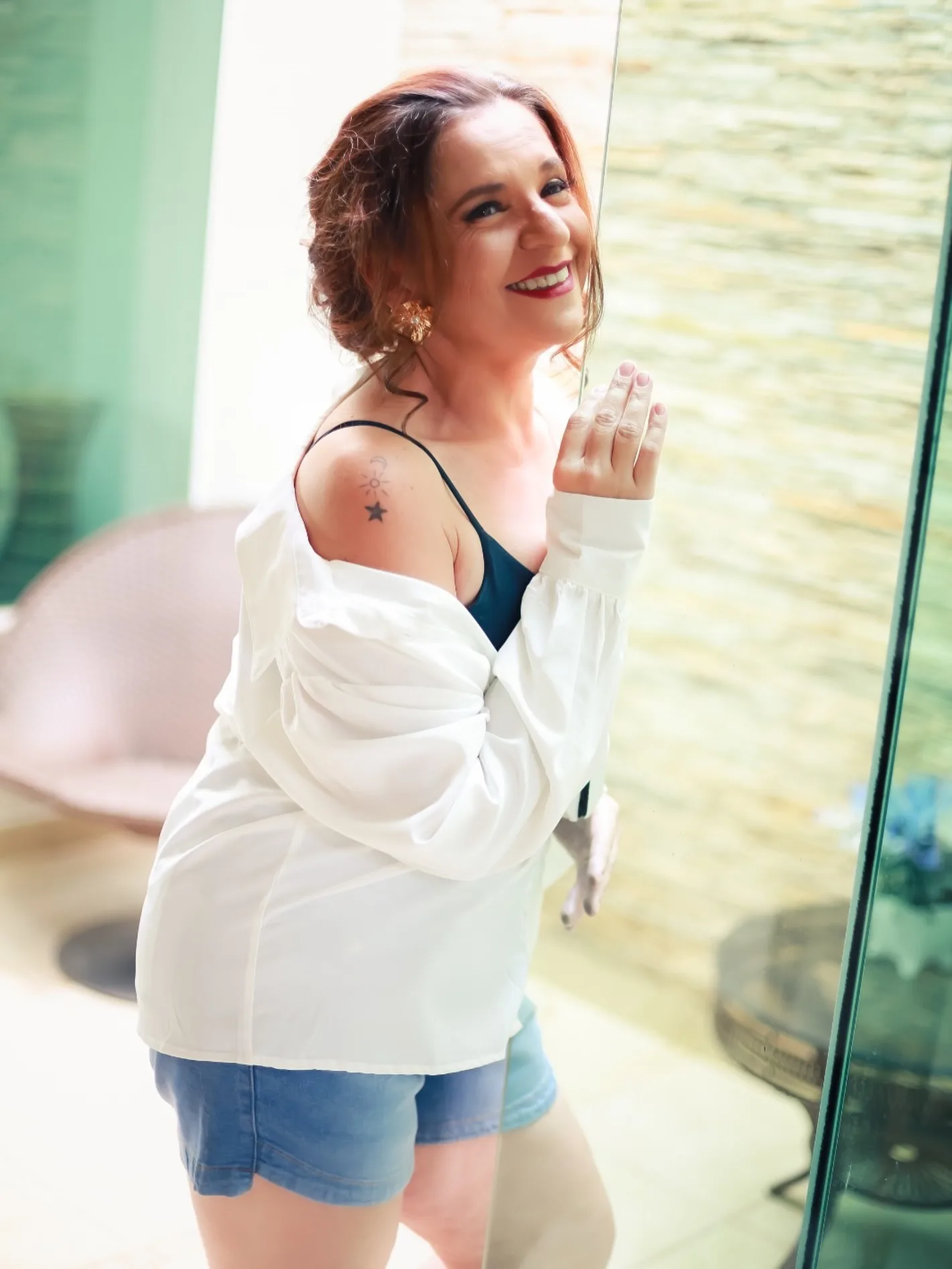 Mulher sorrindo junto à janela, com camisa branca e short jeans