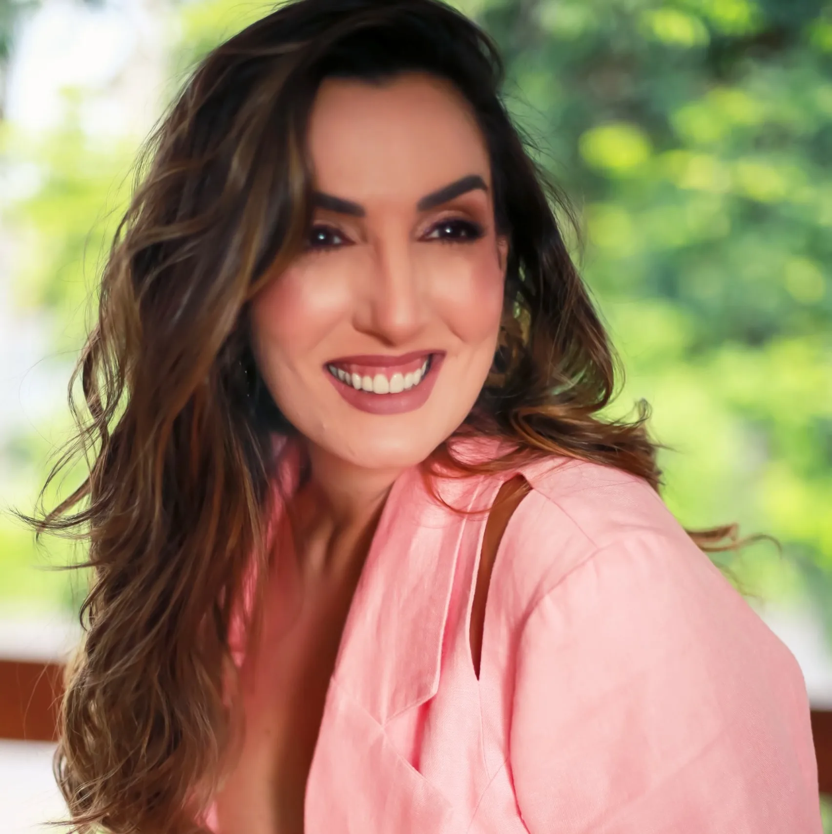 Mulher sorrindo com blusa rosada diante de uma janela