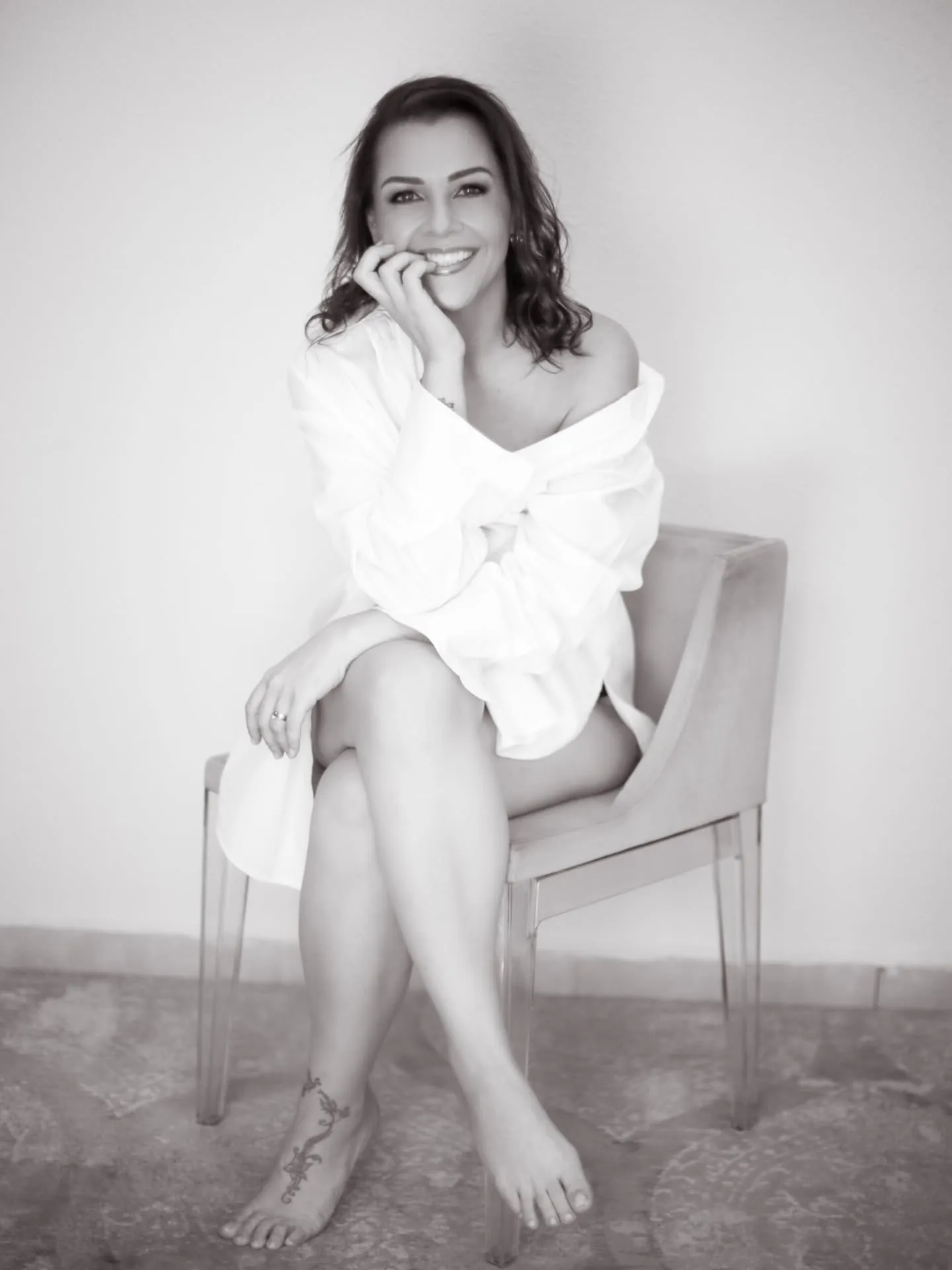 Mulher sorrindo sentada em uma cadeira com camisa branca, em preto e branco